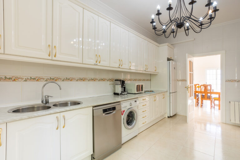 Cocina con gabinetes blancos, grifos dobles, electrodomésticos de acero inoxidable y lámpara de araña negra. Puerta hacia comedor iluminado.