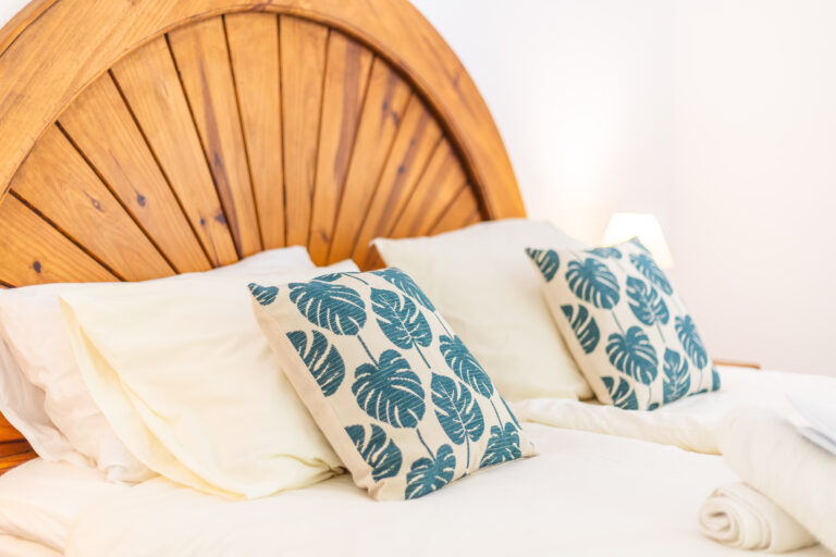 Cabecero de madera arqueada, almohadas blancas con cojines estampados de hojas, cama con sábanas claras y lámpara tenue al fondo.