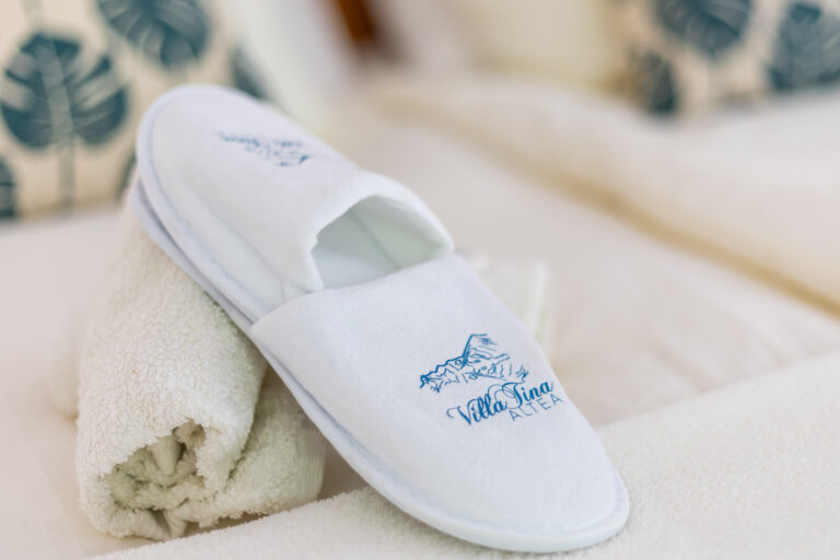 Pantufla blanca de hotel sobre toalla enrollada. Logo azul con texto “Villa Tina Altea”. Fondo difuminado con cojines.