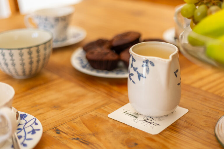 Mesa de madera con tazas de té blancas y azules, jarra, muffins de chocolate y frutero con uvas y plátanos.