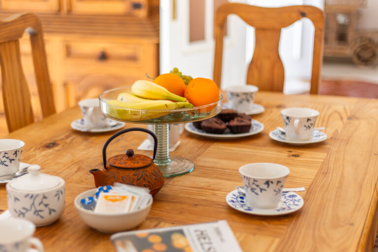 Mesa de desayuno con tetera naranja, cinco tazas, frutero con bananas, naranjas, uvas y platos con magdalenas. Mueble de madera al fondo.