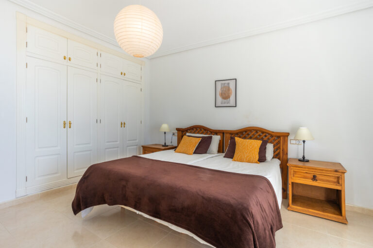 Habitación luminosa con cama grande, colchas marrones, cojines naranjas, lámpara colgante y armario blanco.