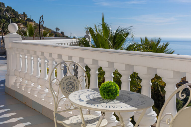 Terraza elegante con barandilla blanca, vistas al mar y palmeras. Mesa y silla de metal blanco con diseño de brújula y bola verde decorativa.