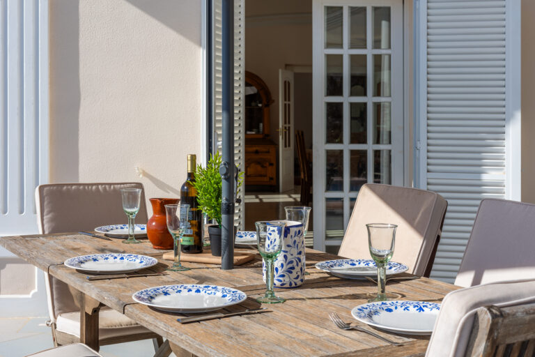Mesa exterior de madera con platos azules, copas de vino, jarra cerámica, botella, jarrón y sillas beige. Puerta al fondo.