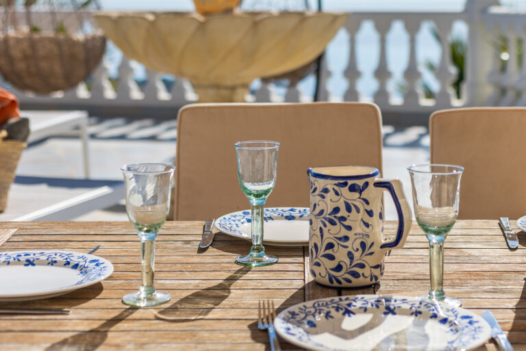 Mesa exterior de madera con platos blancos y azules, copas de vidrio y jarra decorada. Fondo de balaustrada y vegetación.
