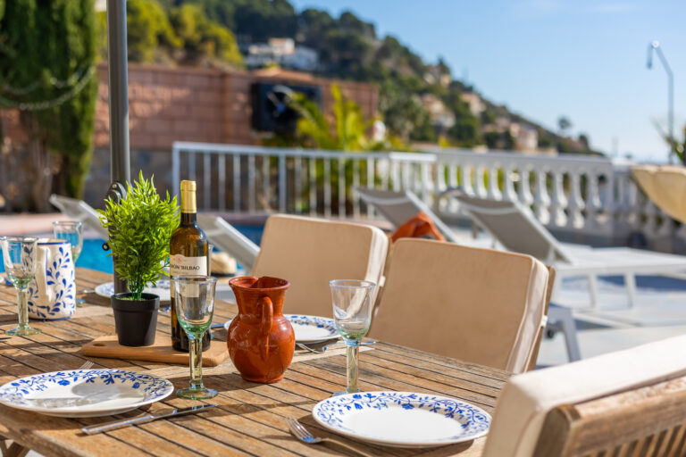 Mesa de madera al aire libre con platos decorados, copas, una planta, jarra, botella de vino y sillas beige. Fondo de piscina y colinas.