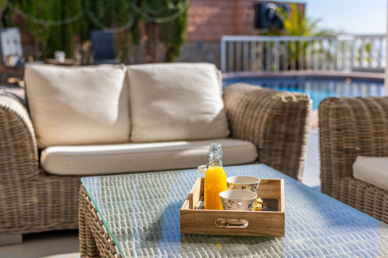 Sofá de mimbre con cojines blancos, mesa de cristal con bandeja de madera, jugo de naranja y tazas, piscina al fondo.