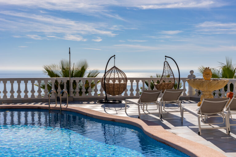 Piscina con agua azul, sillas reclinables, columpios de mimbre, fuente ornamental y vista al mar bajo un cielo despejado.