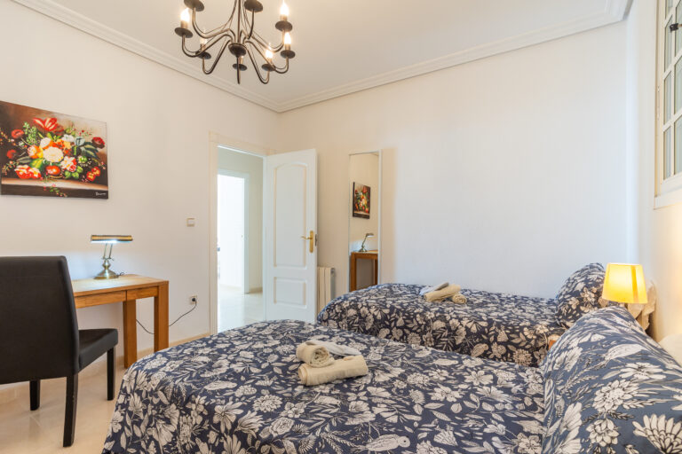 Habitación con dos camas, colchas azul con blanco, escritorio con lámpara, silla negra y lámpara de techo negra. Pared blanca y pintura floral.