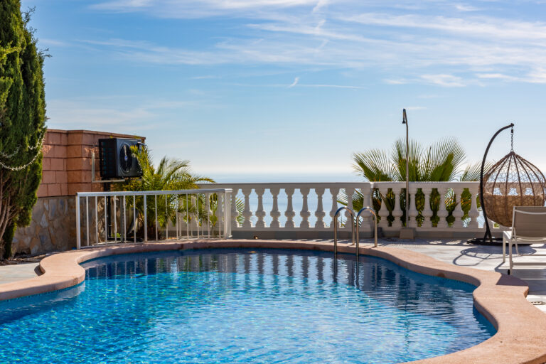 Piscina azul brillante con borde rosa, palmeras verdes y vista al mar. Silla colgante y cielo despejado.