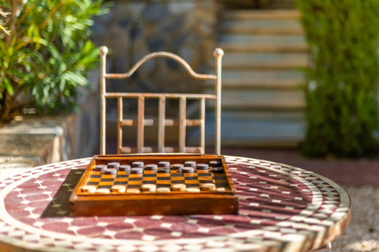 Mesa redonda con diseño geométrico, tablero de ajedrez con fichas de dos colores. Silla de metal y fondo de plantas y escalera.