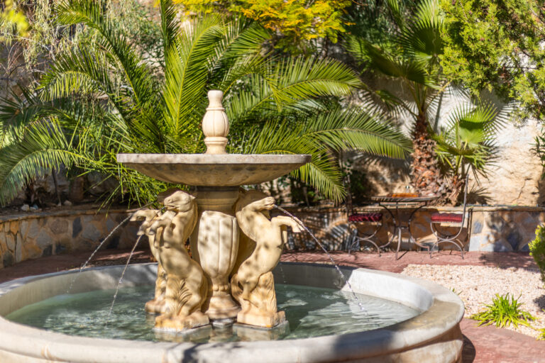 Fuente de piedra con figuras de caballos, palmeras verdes alrededor y muebles de jardín de metal con cojines rojos.