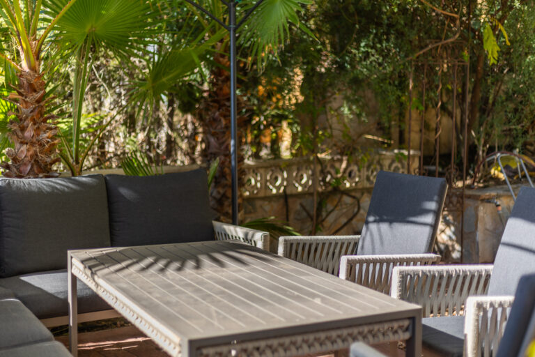 Muebles de exterior con cojines grises, mesa rectangular, rodeados de palmeras y vegetación exuberante en un patio soleado.