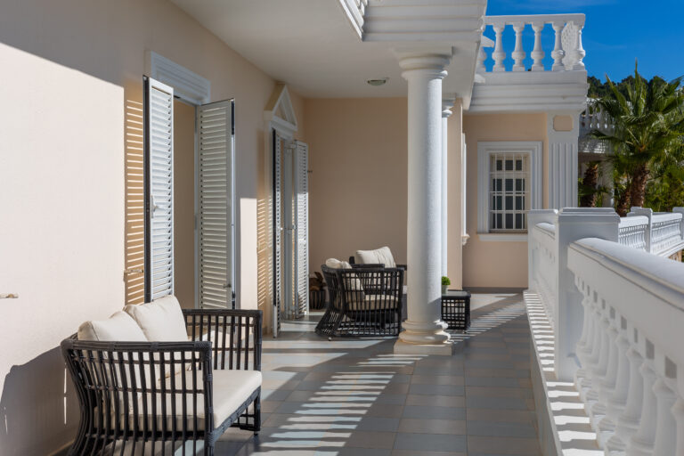 Terraza con columnas blancas, sillones de mimbre negro con cojines claros, puertas tipo persiana y barandilla decorativa.