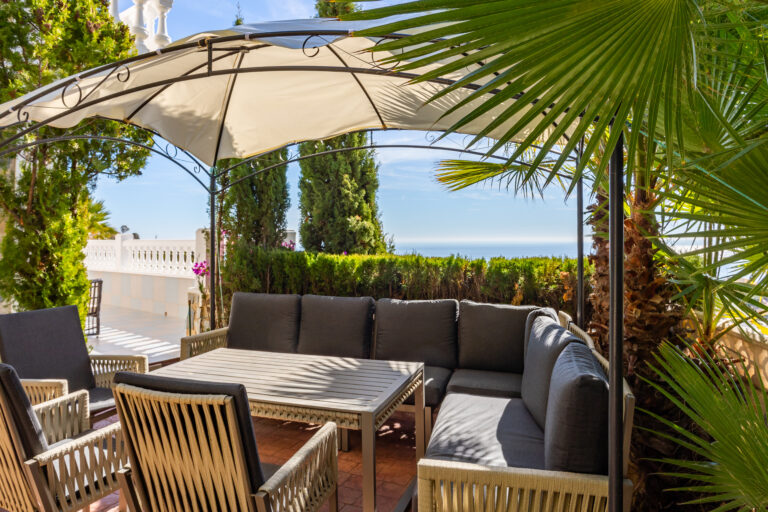 Espacio al aire libre con sofá gris y sillas bajo pérgola, rodeado de palmeras y arbustos; vistas al mar y cielo azul.