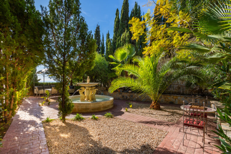 Jardín con fuente central, rodeado de palmeras y cipreses, camino de adoquines rojos y área de estar con sillas y mesa.