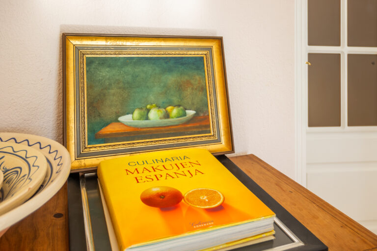 Libro amarillo con texto rojo sobre cocina española frente a un cuadro de frutas verdes en un plato.