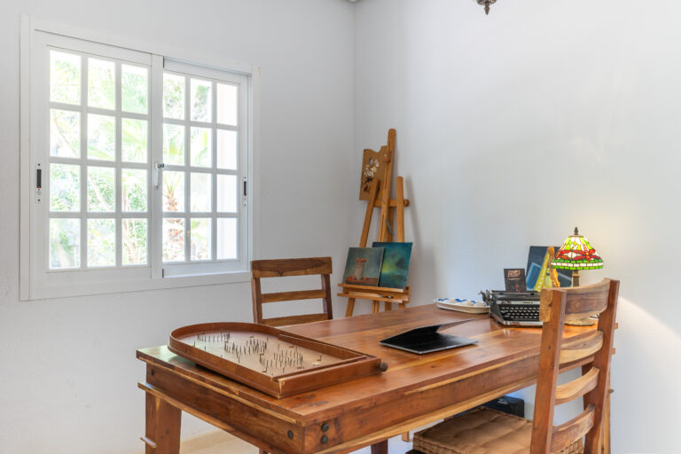 Espacio de trabajo con mesa de madera, lámpara de vidrio, máquina de escribir y pizarra pequeña junto a ventana con vista exterior.