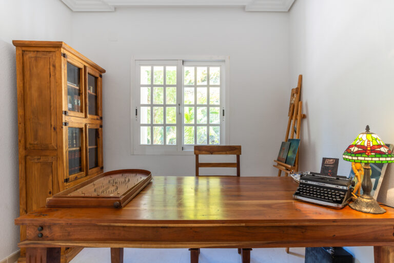 Oficina con muebles de madera, máquina de escribir en escritorio, lámpara de vitral, ventana amplia y mueble con libros.